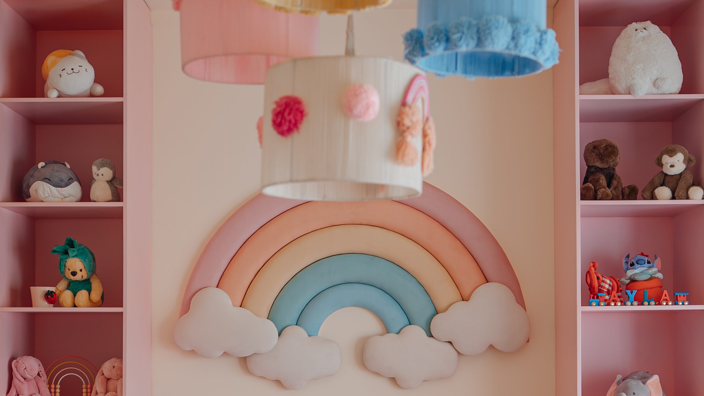 Pastel nursery with a padded rainbow, toy shelves and pompom pendant shades