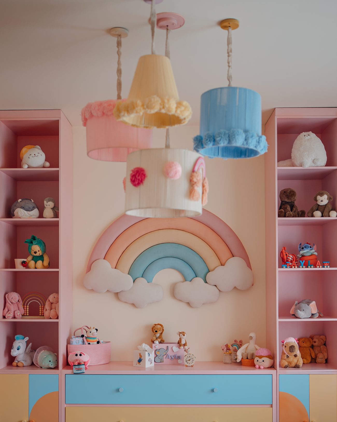 Pastel nursery with pompom pendant shades, a padded rainbow and painted drawers